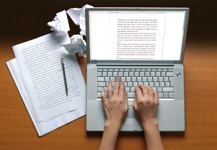 Aufgeklapptes Laptop mit zwei Händen, links liegen mehre beschriebene Papierseiten, ein Stift und Papierknäuel